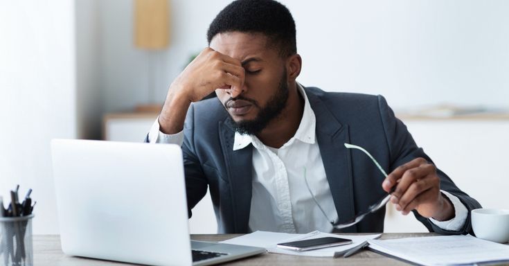 homem negro vestindo paletó e camisa social branca, está com a mão na testa e olhos fechados. O homem está na frente do computador, sentado e estressado com alguma situação. Ele está tentando gerenciar o estresse.